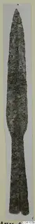 Long corroded metal blade with a pointed tip and tapering tang, photographed vertically on a pale card strip, Arsos