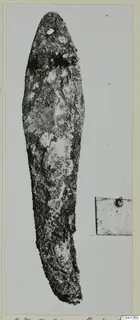 Corroded metal tool with a tapering blade and a small hole near the tip, photographed beside a small label card