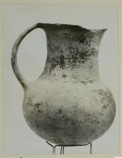 Pottery jug with a single loop handle and a broken rim, standing on a wire stand