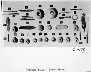 Small finds from Nitovikla T.I laid out in rows: corroded metal pins and rods, a metal ring, round objects with holes and a flat pendant