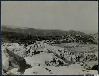 Large limestone blocks and low wall remains in the foreground, with wooded hills and a distant sea beyond, at Vouni.