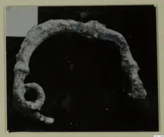 Corroded bronze arc-shaped loop with a round eyelet at its lower left end, photographed against a dark background.