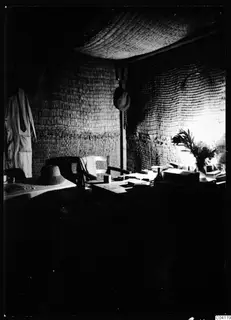 Interior of a bast house at Mersinaki with woven reed walls, a table, a chair and hats.