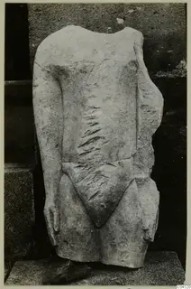 Limestone sculpture of a headless, armless standing figure torso, shown against a dark stone wall.
