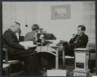 Three men seated in a room in Nicosia, with a table and books; one reads and one smokes a pipe.