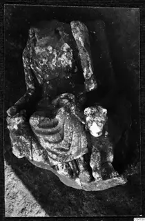 Weathered stone sculpture of a draped figure with an animal head beside it, lying on dark ground at Cholades, Soli.