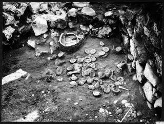Finds in situ at Cholades, Soli: a large broken vessel against a stone wall, with many small round objects scattered on the excavated floor.