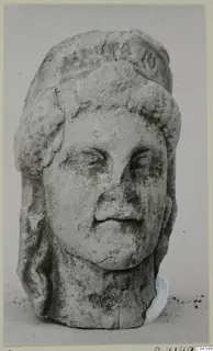 Limestone female head from Soli, shown frontally on a plain background, nose and parts of the headdress broken.
