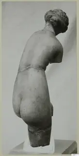 Back view of a marble statue of a nude figure with the head bowed and hair gathered in a coiled bun, the arms broken off.