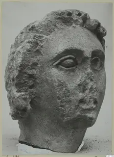 Stone head from Soli, with a large eye, rough surface and broken nose, photographed against a plain background.