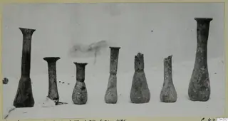 Seven blown glass vessels with narrow necks standing in a row on a pale surface.