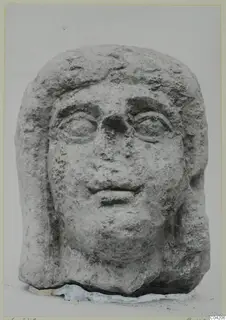 Carved stone head from Soli, frontal view, with hair framing the face and a rough, weathered surface.