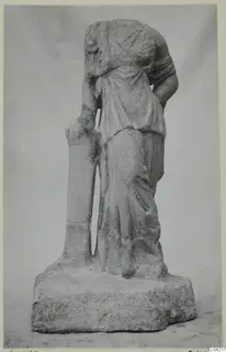 Headless marble statue of a standing draped female figure beside a tall narrow support, on a rough stone base