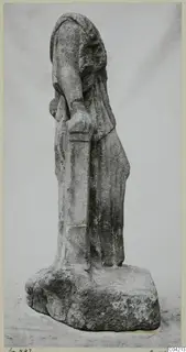 Stone statue of a draped standing figure on a rough block base, photographed against a plain grey backdrop.