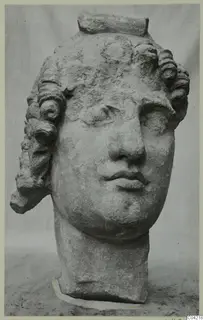 Carved stone head of a figure with curled locks and a raised band across the top, photographed against a grey backdrop. Soli.