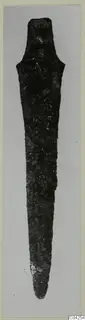 Corroded bronze blade with a short tang, from Milia, photographed upright against a grey background