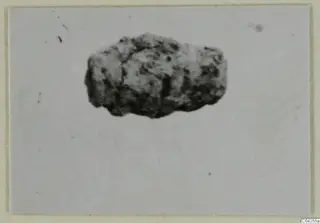 Corroded bronze object from Milia, a small dark lump on a pale mounted print