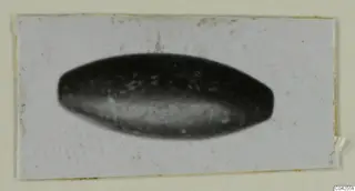 Small dark elongated bead from Milia, photographed on a pale card mount