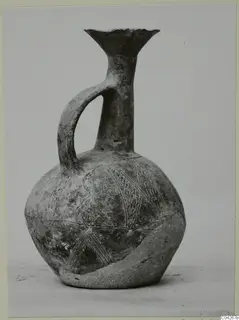 Black-slipped jug with a trefoil-like flared mouth, single strap handle and incised linear decoration, photographed against a grey backdrop.
