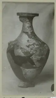 Pale jar with a flat rim, narrow neck and broken body, photographed alone against a plain backdrop.