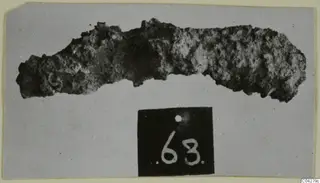 Heavily corroded iron object, elongated, photographed on grey card, from Amathus tombs
