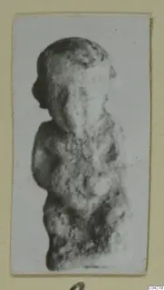 Small worn figure of a standing human figure, seen in side view, in a photograph mounted on a cream card
