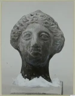 Terracotta head with curled hair, its neck resting on a plain block, photographed against a grey background.