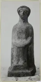 Terracotta figurine of a standing figure with a cap-like headdress and folded arms, on a square base.