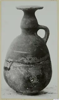 Pottery vessel with a single handle, flaring rim and worn surface, shown against a plain background