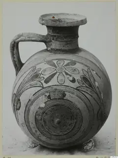 Round-bodied jug with a single handle and painted concentric circles and flower motifs in dark paint on a pale surface.