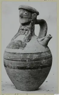Jug with a modelled human face on the neck, a strap handle, a spout and dark painted bands on the body.