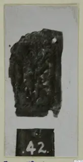 Fragment of tarnished silver sheet with raised scroll-like pattern, shown with a number label below