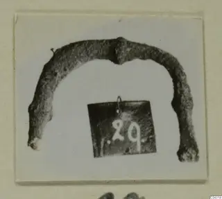 Curved bronze part photographed on a mounted card, with a small number tag hanging below it