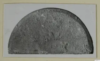 Part of a stone object: a semicircular grey stone fragment with a grainy surface, photographed on a card mount