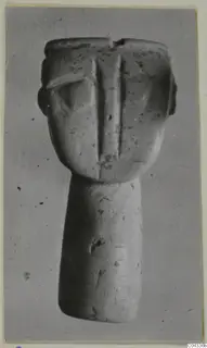 Stone figurine with a flat-topped head, a central ridge down the face and a long cylindrical body, photographed against a grey background.