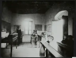 Children seated on wooden benches in a whitewashed village schoolroom with wall maps, and a bearded man standing at the left.