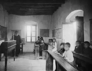 Village school at Ayia Irini: a bearded teacher beside a wall map, with children seated on wooden benches in a whitewashed room.