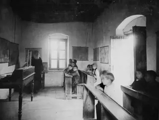 Village school classroom at Ayia Irini with a bearded teacher standing and children seated on wooden benches