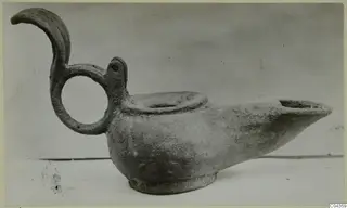 Metal oil lamp with a ring handle topped by a curled projection, spout to the right, shown in side view.