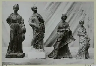 Four standing female terracotta figurines in draped robes, set in a row on a table against a draped cloth.