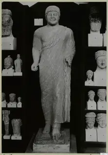 Standing limestone statue of a draped male figure, displayed on a base between shelves of heads and busts