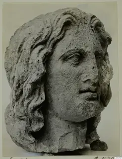 Carved stone male head with long wavy hair, shown in three-quarter view against a plain background.
