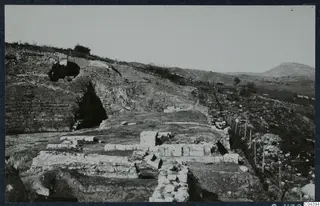 The theatre at Soli seen from the east, with excavated stone blocks and steps in the foreground and hills behind.