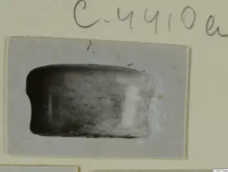 Small photograph of a dark stone seal or gem-like object, roughly rectangular with rounded ends, mounted on a card