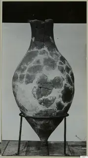 Large bulbous vessel with a narrow neck and broken rim, standing on a metal ring stand against a plain backdrop