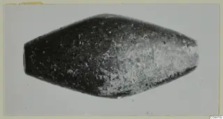 Elongated double-cone find with a dark, mottled surface, photographed on a grey background