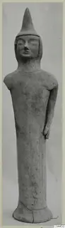 Standing terracotta figure with a tall pointed headdress and elongated body, photographed against a grey backdrop.