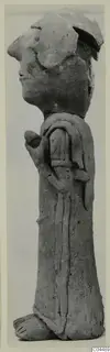 Side view of a terracotta standing figurine with a flat headdress and one arm bent across its body.