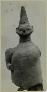 Terracotta figurine from Ayia Irini of a standing human figure with a tall conical head and one arm bent at the side.