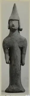 Terracotta figurine of a standing figure with a tall conical head covering and arms held at the sides.